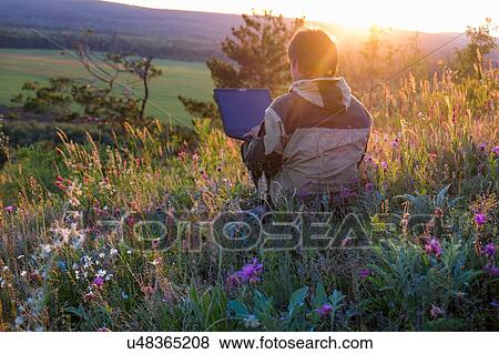 Stock Photo - Mid adult man sitting in wildflower meadow using laptop. Fotosearch