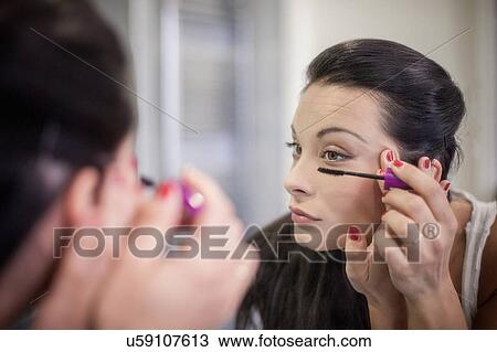 Stock Image - Over the shoulder mirror image of young woman applying mascara. Fotosearch