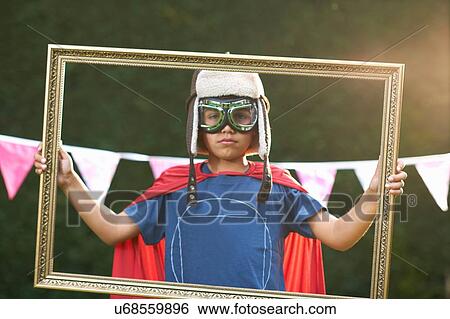 Stock Photograph - Portrait of boy looking through picture frame wearing cape, goggles and flying hat. Fotosearch
