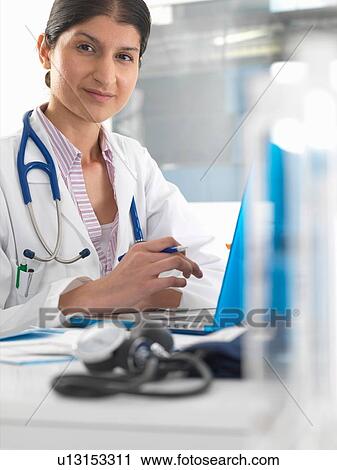 Stock Image - Portrait of female doctor at desk working on laptop. Fotosearch