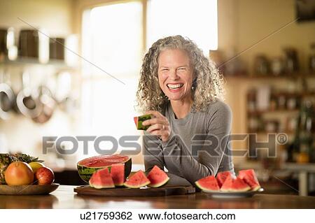 Stock Image - Portrait of mature woman eating watermelon in kitchen. Fotosearch