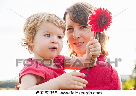 Stock Photography - Portrait of mid adult mother and toddler daughter holding red flower in field. Fotosearch