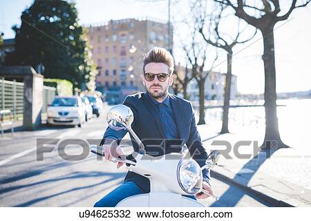 Stock Image - Portrait of stylish young man sitting on moped at roadside. Fotosearch