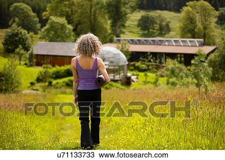 Stock Image - Rear view of mature woman carrying yoga mat walking in eco lodge field. Fotosearch