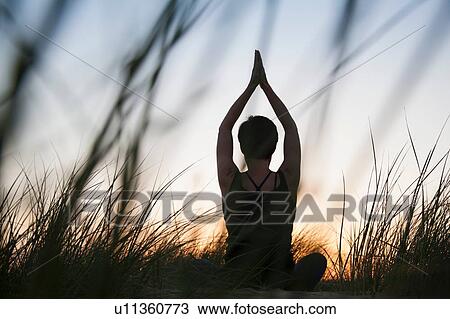 Stock Image - Rear view of mid adult woman practicing yoga in silhouetted long grasses at sunset. Fotosearch