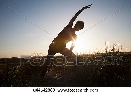 Stock Photo - Rear view of mid adult woman practicing yoga in silhouetted long grass at sunset. Fotosearch