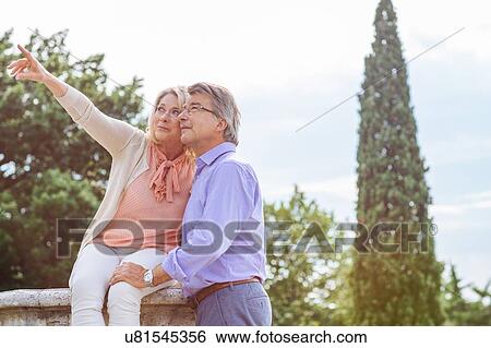 Stock Photograph - Senior couple sitting on wall, looking at view. Fotosearch
