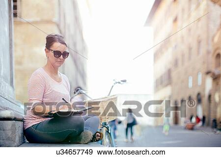 Stock Photo - Side view of mid adult woman wearing sunglasses sitting crossed legged writing in notebook. Fotosearch