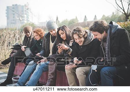 Stock Image - Six young adult friends networking on smartphones and digital tablets in park. Fotosearch
