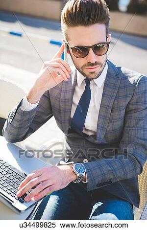 Stock Photography - Stylish young businessman using smartphone and laptop at sidewalk cafe. Fotosearch