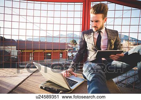Stylish young man sitting on footbridge using laptop and digital tablet View Large Photo Image Stock Image - Stylish young man sitting on footbridge using laptop and digital tablet. Fotosearch