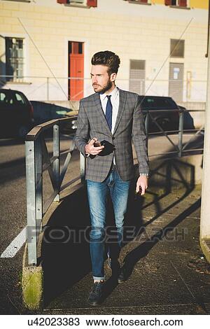 Stock Image - Stylish young man with smartphone walking on sidewalk. Fotosearch