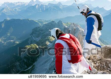 Stock Photography - Two male BASE jumpers standing on edge of mountain looking down, Dolomites, Italy. Fotosearch