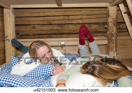 Stock Image - Two young women lying on bed in log cabin chatting. Fotosearch