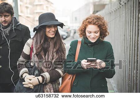 Two young women strolling along street reading smartphone texts View Large Photo Image Stock Image - Two young women strolling along street reading smartphone texts. Fotosearch