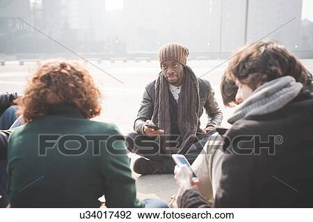 Stock Image - Young adult friends sitting networking on smartphones. Fotosearch