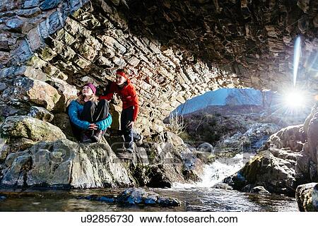 Stock Image - Young couple hiking, resting under bridge, Ashness Bridge, Keswick, Lake District, Cumbria, United Kingdom. Fotosearch