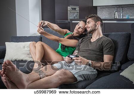 Stock Photo - Young couple on sofa taking smartphone selfie. Fotosearch