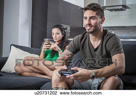 Stock Image - Young couple on sofa using smartphone and gaming control. Fotosearch