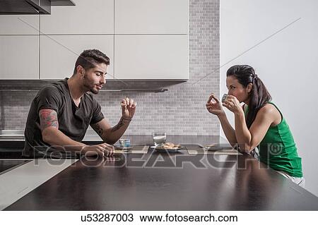 Stock Image - Young couple opposite each other having breakfast in kitchen. Fotosearch