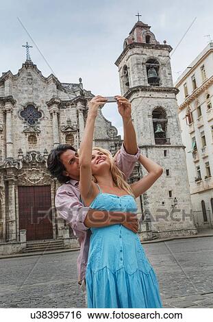 Stock Photograph - Young couple taking selfie with smartphone in the Plaza de la Cathedral of Havana, Cuba. Fotosearch