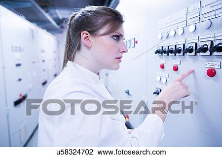 Stock Image - Young female scientist pointing at machine in technical room. Fotosearch