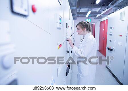 Stock Image - Young female scientist turning switch on machine in technical room. Fotosearch