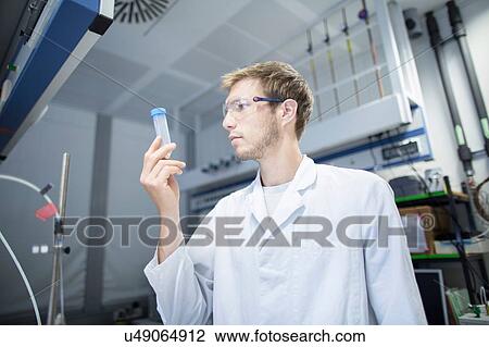 Stock Image - Young male scientist looking at sample bottle in lab. Fotosearch