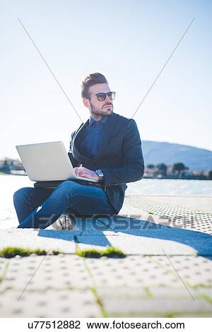 Stock Image - Young man looking over his shoulder whilst using laptop on lakeside step. Fotosearch