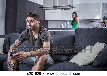 Young man on sofa using gaming control watched by angry girlfriend View Large Photo Image Picture - Young man on sofa using gaming control watched by angry girlfriend. Fotosearch