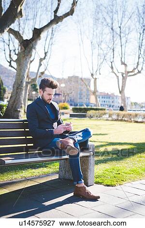 Stock Photo - Young man sitting on park bench reading smartphone texts. Fotosearch