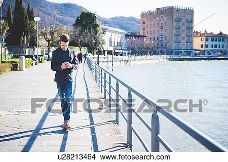 Picture - Young man strolling and reading smartphone texts on lakeside, Rovato, Brescia, Italy. Fotosearch