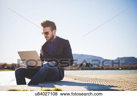 Stock Photo - Young man using laptop on lakeside step. Fotosearch