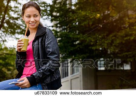 Stock Photo - Young woman in park holding smartphone and drinking smoothie. Fotosearch