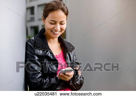 Stock Image - Young woman leaning against pillar reading smartphone texts. Fotosearch