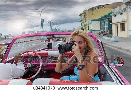 Stock Image - Young woman photographing from a vintage car on the Havana' Malecon, Cuba. Fotosearch