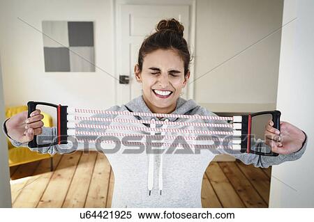 Young woman straining to exercise using chest expander in living room View Large Photo Image Stock Photography - Young woman straining to exercise using chest expander in living room. Fotosearch