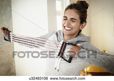 Young woman straining to exercise with chest expander in living room View Large Photo Image Stock Photo - Young woman straining to exercise with chest expander in living room. Fotosearch