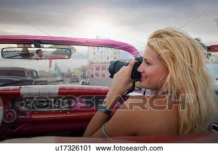 Stock Image - Young woman taking pictures from a vintage car on the Havana' Malecon, Cuba. Fotosearch