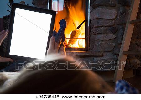 Stock Photo - Young woman using digital tablet with feet up in front of fireplace. Fotosearch