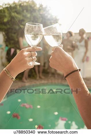 Stock Image - Arms of two people holding wine glasses making toast in front of swimming pool. Fotosearch