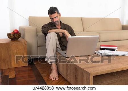 Stock Photo - "Barefoot mature man sitting on wooden floor with laptop, looking down". Fotosearch