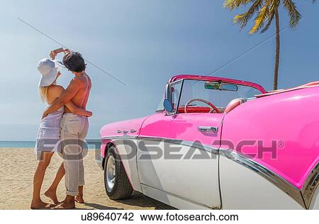 Stock Image - Couple taking selfie with smartphone beside Convertible on beach. Fotosearch