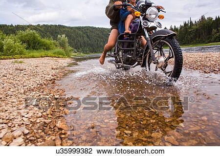 Stock Image - "Father and son on motorbike in rural setting, riding through stream, low section". Fotosearch