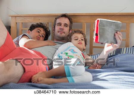 Stock Image - "Father and two sons relaxing on bed, looking at digital tablet". Fotosearch