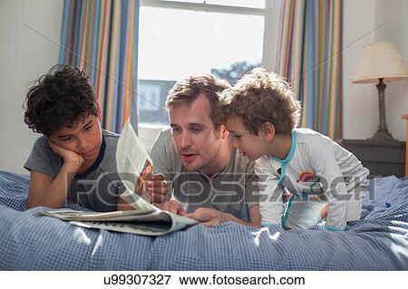Stock Photo - "Father and two sons relaxing on bed, looking at newspaper". Fotosearch