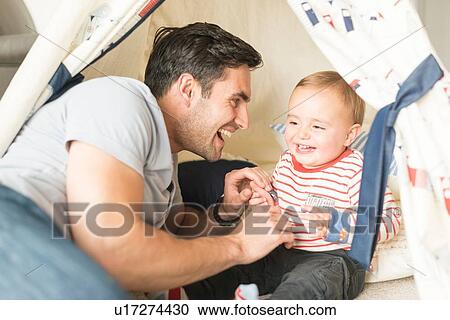 Stock Image - "Father and young son sitting in play tent indoors, laughing together". Fotosearch