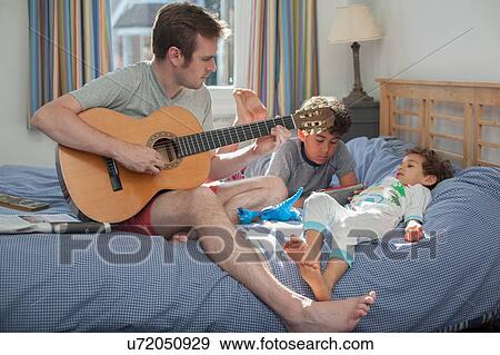 Father playing guitar while two sons relax on bed View Large Photo Image Stock Photo - Father playing guitar while two sons relax on bed. Fotosearch