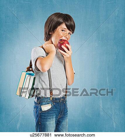 Stock Image - "Freckled boy wearing braces, carrying bundle of books over shoulder, looking at camera, biting apple". Fotosearch