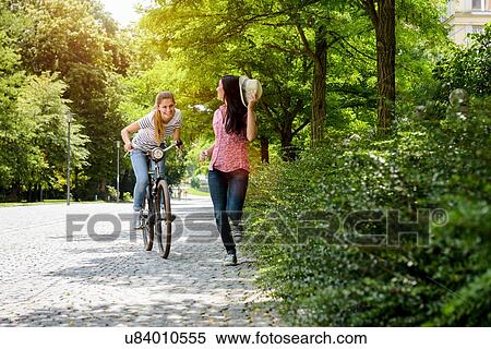 Stock Photography - Front view of young smiling woman on bicycle chasing young woman holding panama hat. Fotosearch